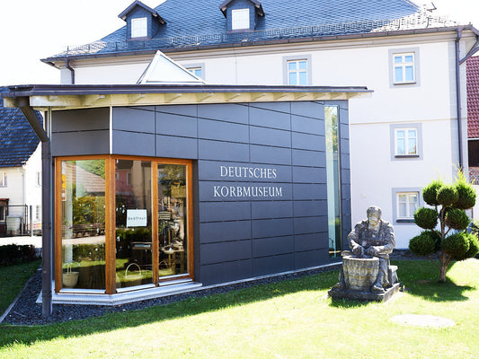 German Basket Museum exterior with modern entrance facade and seated basketmaker statue in front garden / ドイツかご博物館の外観、モダンな入口ファサードと前庭に座るかご職人の像