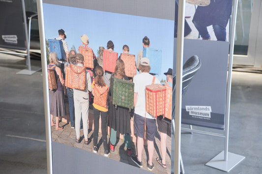 Photograph of an exhibition panel showing people standing in a row wearing woven basket backpacks, displayed at the Swedish exhibition of the World Wicker and Weaving Festival 2023 / 世界かご編み大会2023スウェーデン展示で紹介されている、背負い籠を身につけた人々の写真展示を撮影した様子